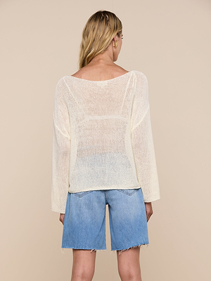 Woman faces away from the camera in a relaxed fit, sheer Ethan Knit Pullover with an off-shoulder neckline, paired with light blue frayed hem denim shorts, against a plain beige background.