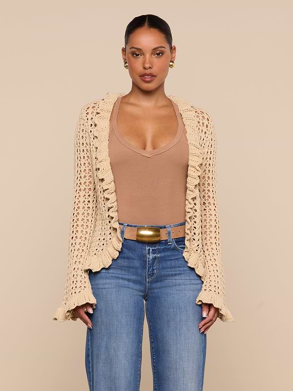 A woman stands against a plain white background, layering the Solaris Crocheted Cardigan—a beige open-stitch crochet design—over a tan top, blue jeans, and a wide belt.