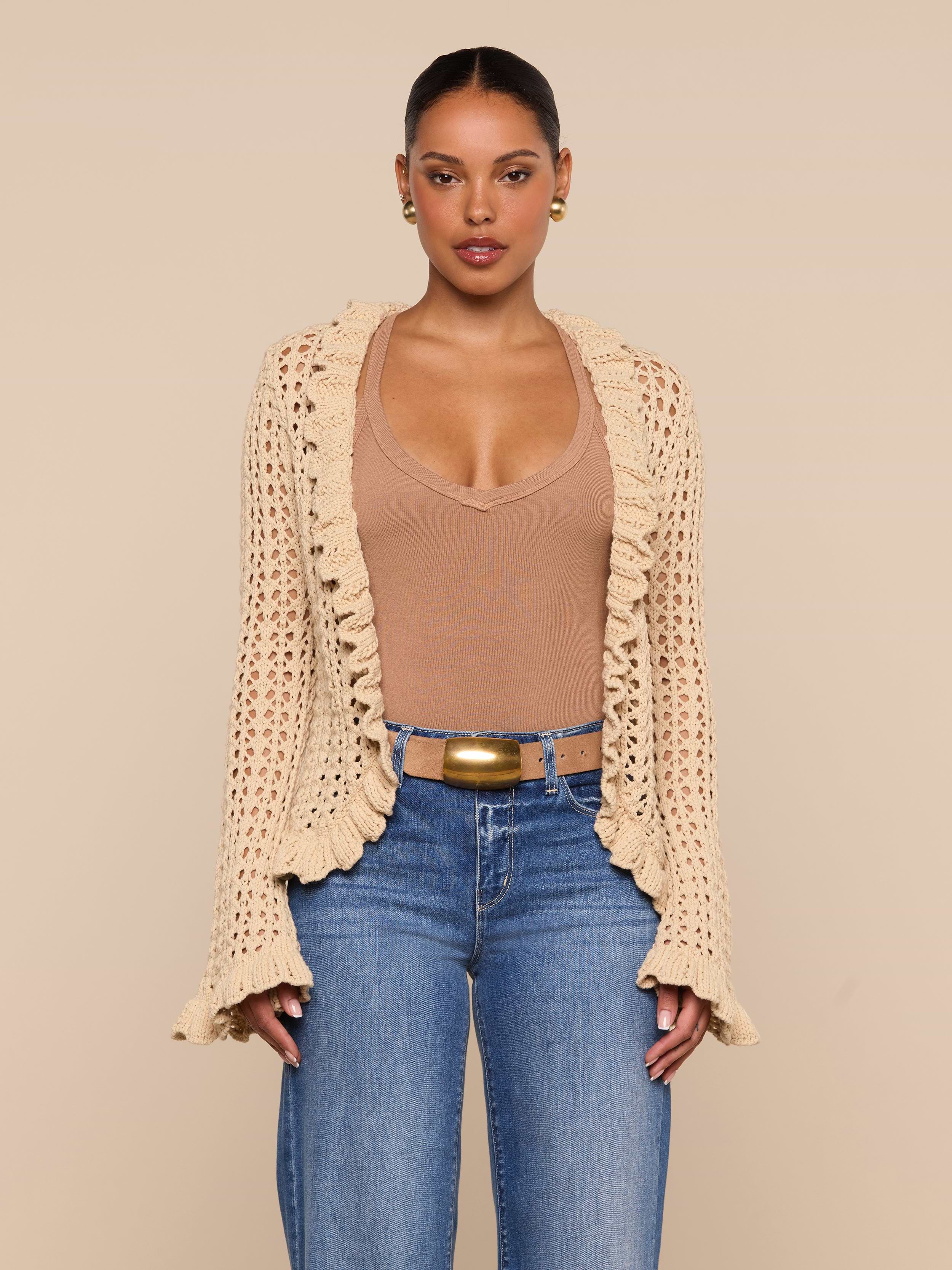 A woman stands against a plain white background, layering the Solaris Crocheted Cardigan—a beige open-stitch crochet design—over a tan top, blue jeans, and a wide belt.