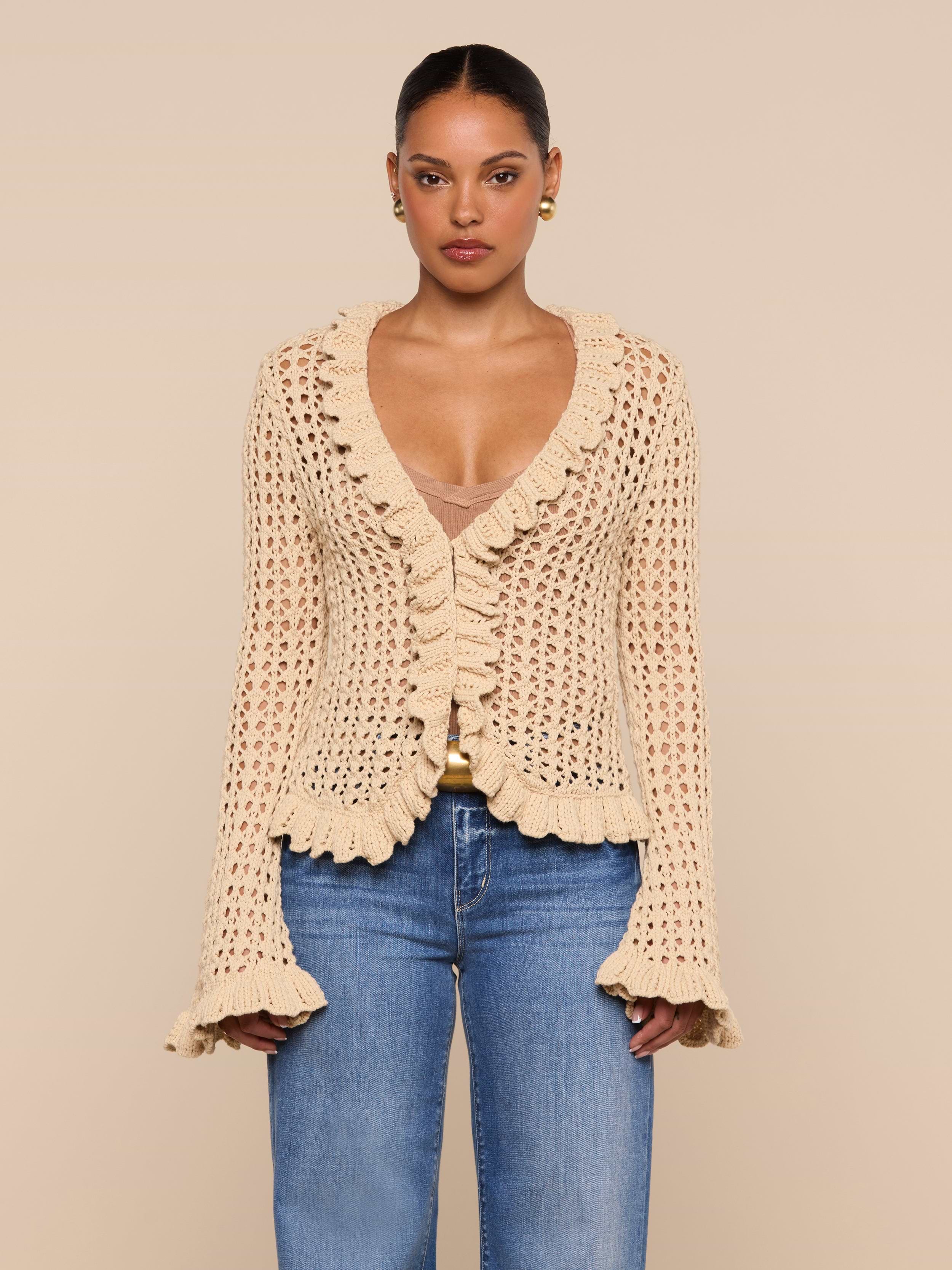 A woman is wearing the Solaris Crocheted Cardigan—a beige, ruffled, open-stitch crochet design with bell sleeves—paired with blue jeans and standing against a plain light background.