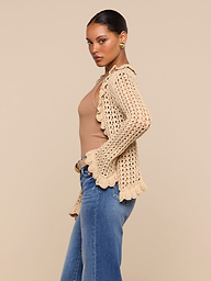 A woman stands sideways against an off-white background, wearing the Solaris Crocheted Cardigan in tan over a beige top and blue jeans, with her hair pulled back and accessorized with gold earrings.