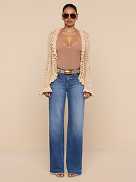 A woman stands facing forward against a plain white background, wearing sunglasses, the Solaris Crocheted Cardigan with open-stitch details in beige, a tan deep-neckline top, blue wide-leg jeans, and brown heels.