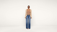 A woman wearing sunglasses, a beige top with a matching open-knit cardigan, blue high-waisted wide-leg jeans, and sandals stands against a plain white background.