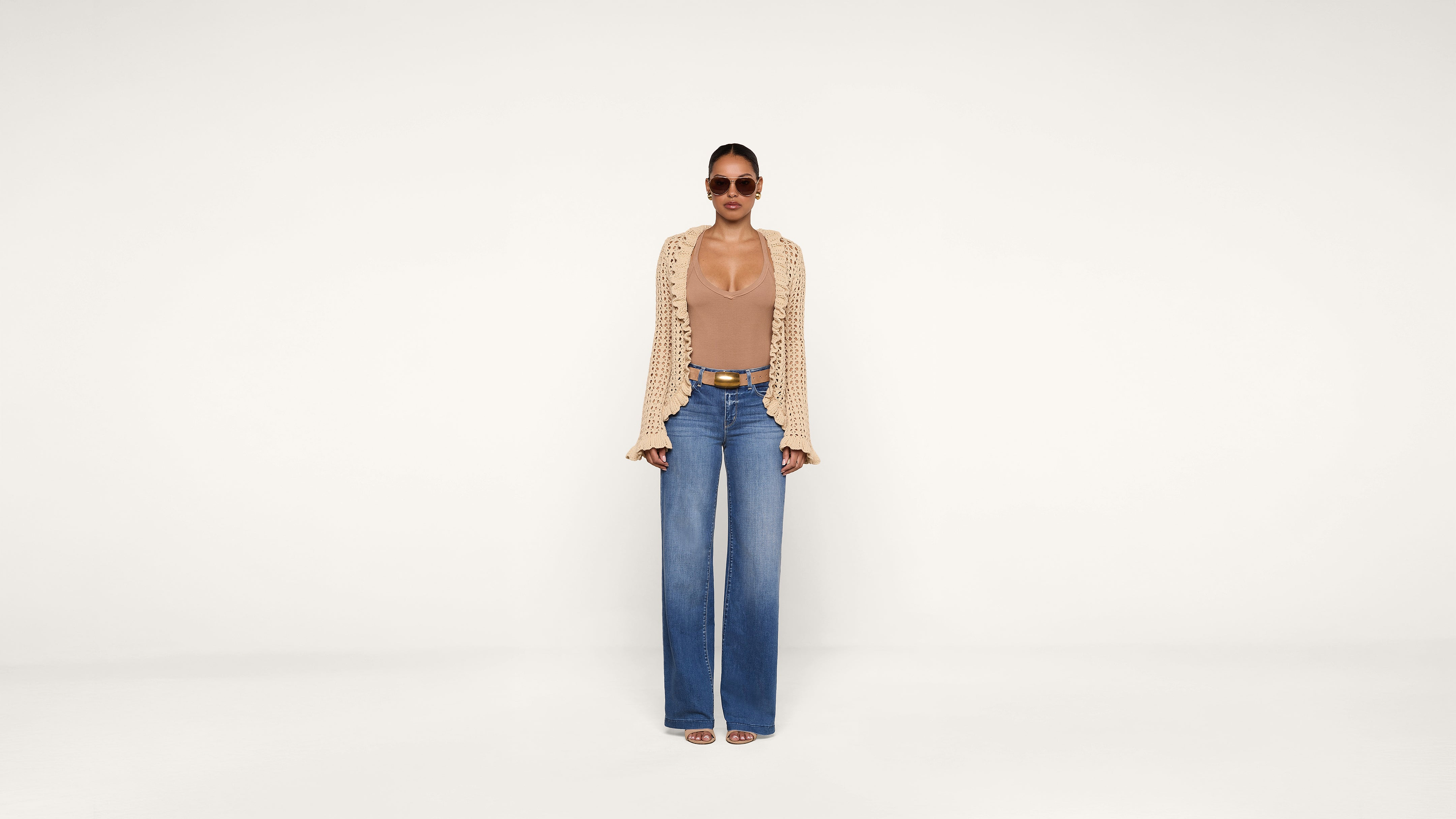 A woman wearing sunglasses, a beige top with a matching open-knit cardigan, blue high-waisted wide-leg jeans, and sandals stands against a plain white background.