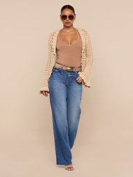 A woman poses confidently against a plain white background, wearing sunglasses, a tan top, the Solaris Crocheted Cardigan as a lightweight layer, blue wide-leg jeans, and sandal heels.
