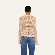 A person stands facing away, wearing the Solaris Crocheted Cardigan—a beige open-stitch, long-sleeve top with scalloped edges—paired with blue jeans. Their hair is styled in a neat bun against a plain, light-colored background.