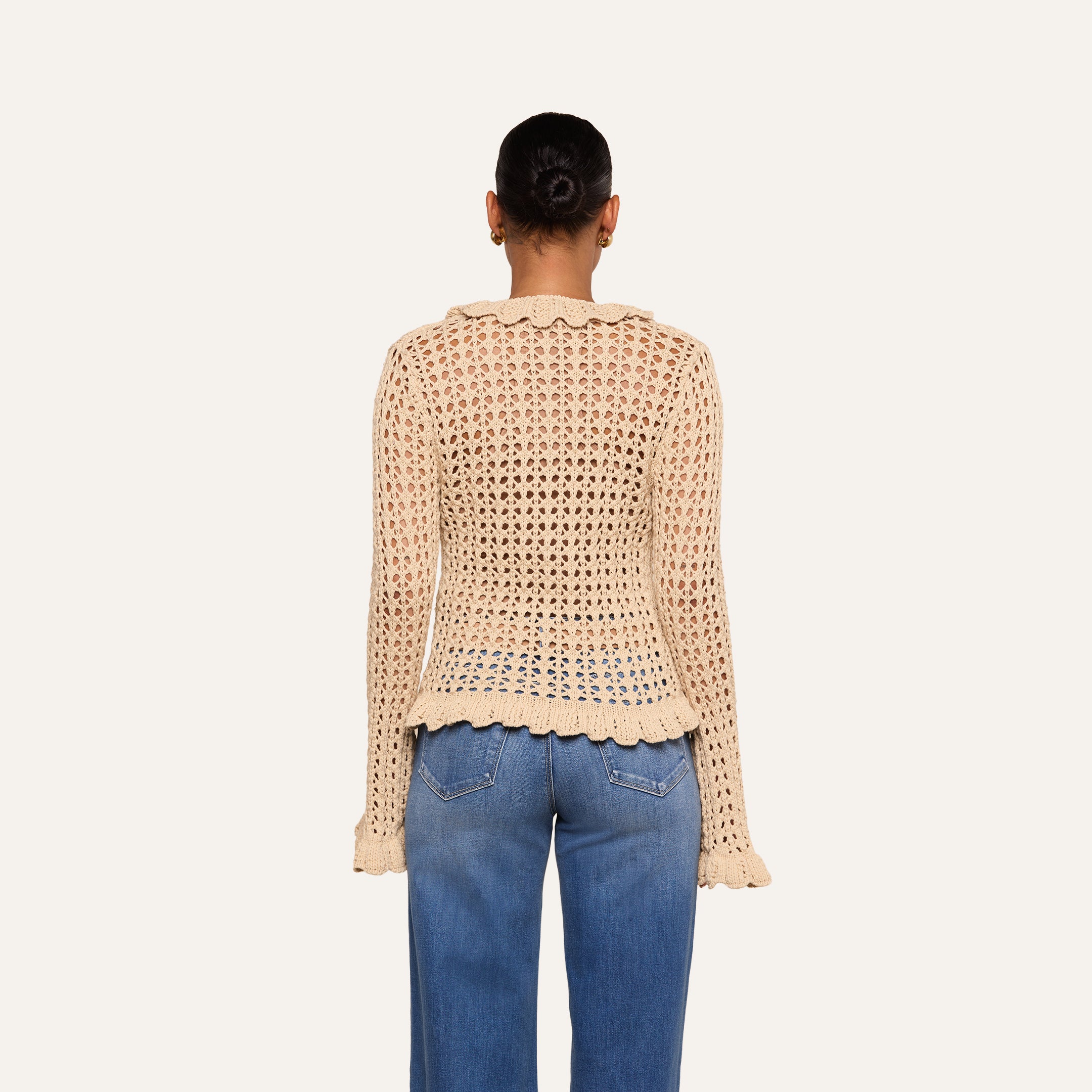A person stands facing away, wearing the Solaris Crocheted Cardigan—a beige open-stitch, long-sleeve top with scalloped edges—paired with blue jeans. Their hair is styled in a neat bun against a plain, light-colored background.