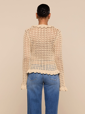 A person, facing away, wears the Solaris Crocheted Cardigan in beige with open-stitch design and scalloped hem and cuffs—an ideal lightweight layer—paired with high-waisted blue jeans against a plain light beige background.
