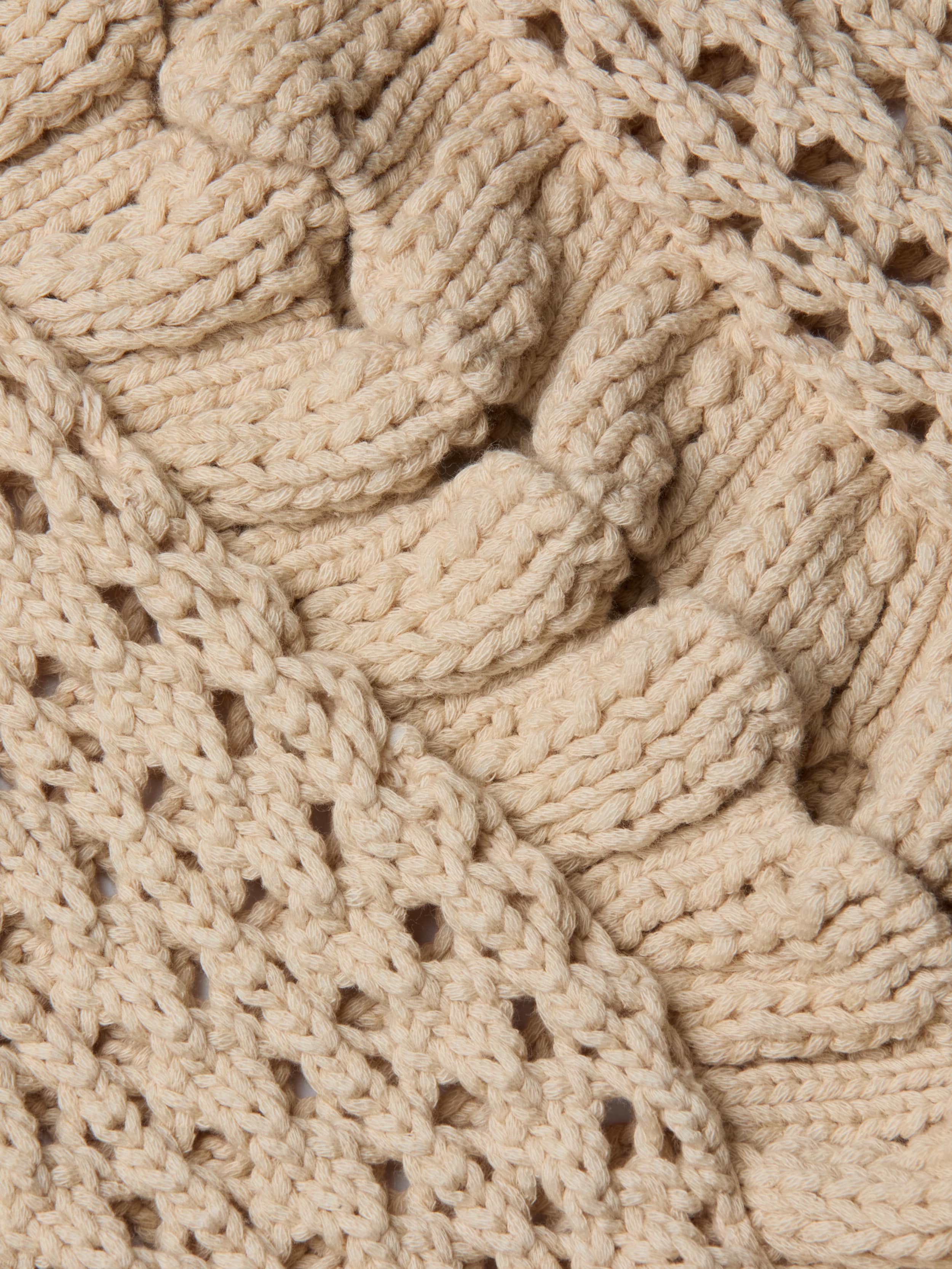 Close-up of the Solaris Crocheted Cardigan in beige reveals its intricate, chunky, and textured knit with ribbed and open-stitch sections—perfect for a lightweight layer.