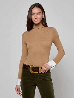 A woman with long brown hair wears the Flora Turtleneck by L'AGENCE, styled with dark green pants and a wide brown belt with a gold buckle, standing against a plain light gray background.