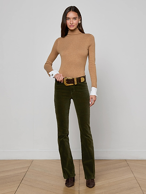 A woman with long hair stands indoors against a white wall, wearing the L'AGENCE Flora Turtleneck in tan, olive green belted pants, and brown shoes. She has a neutral expression.