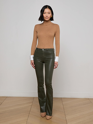 A woman with short black hair and a neutral expression stands indoors against a white wall, wearing the L'AGENCE Flora Turtleneck with French cuffs, dark green leather pants, and beige pointed-toe shoes.