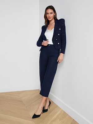 A woman stands against a white wall on a wooden floor, wearing the Bethany Knit Blazer in midnight blue with silver buttons, paired with a white top, navy cropped pants, and black pointed heels.