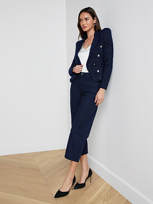 A woman with long dark hair stands in a corner, wearing the Bethany Knit Blazer with gold buttons, a white top, navy cropped pants, and black heels. She rests one hand on her blazer and looks slightly to the side.