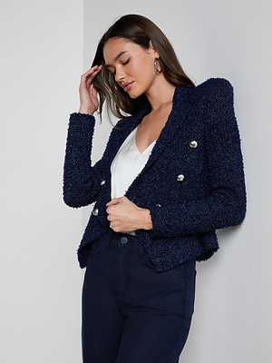 A woman stands against a white wall in the Bethany Knit Blazer—midnight blue with silver buttons—paired with a white top and dark jeans. With long brown hair, hoop earrings, and eyes closed, she touches her forehead gently.