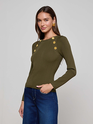 A woman with long brown hair wears the L'AGENCE Gretchen Pullover—a fitted olive-green sweater with decorative shoulder buttons—paired with dark blue jeans, standing against a plain light background with one hand in her pocket.