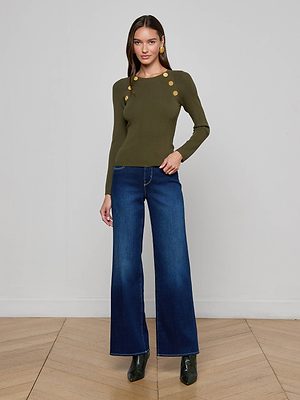 A woman with straight brown hair poses confidently indoors against a plain gray wall, wearing the fitted Gretchen Pullover by L’AGENCE in deep olive with gold shoulder buttons, styled with wide-leg blue jeans and black ankle boots.