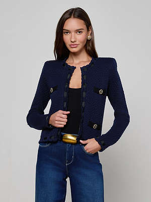 A woman with long dark hair wears the L'AGENCE Astor Cardigan in midnight blue over a black top and blue jeans with a gold belt, standing against a light gray background and smiling slightly, facing forward.