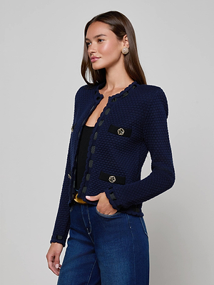 A woman with long brown hair models the L'AGENCE Astor Cardigan in midnight blue with black trim and buttons, paired with a black top and blue jeans, exuding Parisian-inspired elegance against a light gray backdrop.