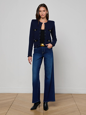 A woman poses indoors on a wooden floor against a light wall, exuding Parisian-inspired elegance in the midnight blue Astor Cardigan by L'AGENCE, paired with a black top, wide-leg blue jeans, a gold belt, and black heeled boots.