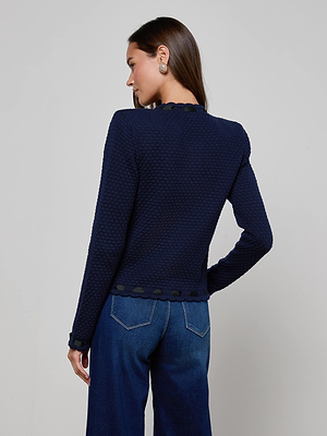 A woman stands facing away, highlighting the back of the L'AGENCE Astor Cardigan in textured midnight blue with scalloped edges. She pairs it with wide-leg blue jeans and shows off a statement earring with her straight brown hair tucked behind her ear.