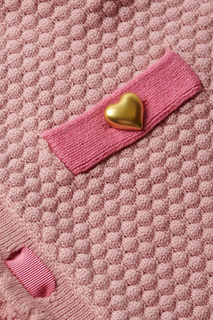 Close-up of the pink textured fabric on the L'AGENCE Astor Cardigan, highlighting a rectangular pink ribbon with a shiny gold heart-shaped button and another partially visible ribbon at the corner.