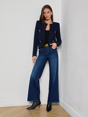 A woman with long brown hair leans against a white wall, radiating Parisian-inspired style in a midnight blue L'AGENCE Astor Cardigan with gold buttons, paired with a black top, wide-leg jeans, yellow belt, and black ankle boots.