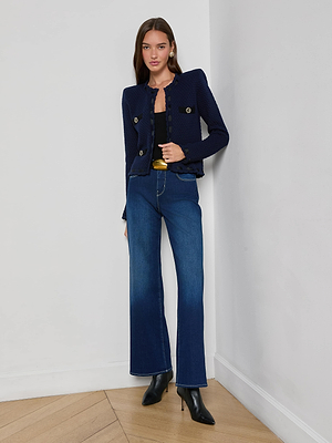 A woman with straight brown hair and a neutral expression stands indoors against a white wall, wearing the L'AGENCE Astor Cardigan—a midnight blue textured jacket—paired with a black top, flared blue jeans, and black heeled boots.