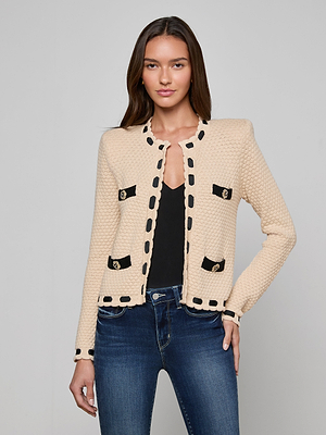 A woman models the L'AGENCE Astor Cardigan, a cream textured knit with black trim and decorative buttons, layered over a black top and blue jeans against a gray background.