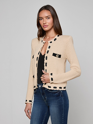 A woman with long brown hair models the L'AGENCE Astor Cardigan, a cream textured knit jacket with black trim and buttons, worn over a black top and blue jeans against a plain light gray background for Parisian-inspired style.