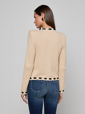 A woman with long brown hair faces away, wearing the L'AGENCE Astor Cardigan—a cream knit piece with black circular trim on the collar, cuffs, and hem—paired with blue jeans for a Parisian-inspired look against a light gray backdrop.