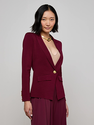 A woman with medium-length black hair wears the L'AGENCE Chamberlain Knit Blazer in maroon with gold buttons over a beige top and burgundy pants, accessorized with a gold necklace, standing against a plain light gray background.