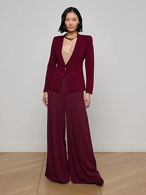 A woman poses against a plain wall in the L'AGENCE Chamberlain Knit Blazer with wide-leg trousers in deep burgundy, styled with a nude top and black choker. The wooden floor complements her modern, elegant look for a striking effect.