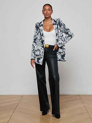 A woman poses confidently indoors on a wooden floor, wearing L'AGENCE's Eileen Jacquard Knit Blazer in an oversized fit, styled with a white top, black wide-leg pants with a gold-buckled belt, and black pointed shoes against a light wall.