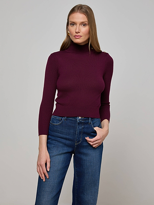 A woman with long straight hair wears the L'AGENCE Kamdyn Cropped Sweater, a fitted maroon long-sleeve mock neck in lightweight knit, paired with blue jeans, standing against a plain light gray background.