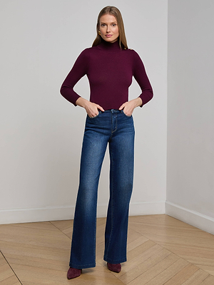 A woman with straight hair poses indoors against a plain wall, wearing the L'AGENCE Kamdyn Cropped Sweater in maroon, high-waisted flared blue jeans, and maroon pointed-toe shoes, with her hands in her pockets.