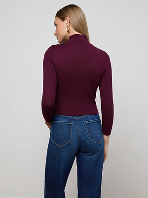 A woman with straight, light brown hair faces away, wearing the L'AGENCE Kamdyn Cropped Sweater in burgundy with blue high-waisted jeans and gold hoop earrings. The background is a plain light gray.