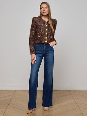 A woman with long straight hair stands indoors against a plain wall, wearing the brown Una Knit Cardigan, high-waisted blue jeans, and beige heels. Her left hand rests in her pocket.