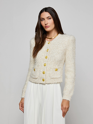 A woman with long brown hair wears the Una Knit Cardigan with gold buttons and white wide-leg pants, standing against a plain light gray background.