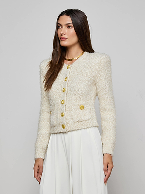 A woman with long brown hair models the Una Knit Cardigan, a cream eyelash knit with gold buttons, paired with a white skirt against a light gray background.