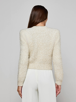 A woman with long brown hair stands facing away from the camera, wearing the Una Knit Cardigan with gold buttons and white pants, set against a plain light background.