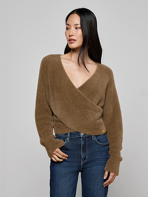 Woman wearing the Antonella Crossover Sweater—a cozy, chic brown fuzzy wrap-style V-neck—paired with blue jeans, standing against a plain light gray background and looking to the side.