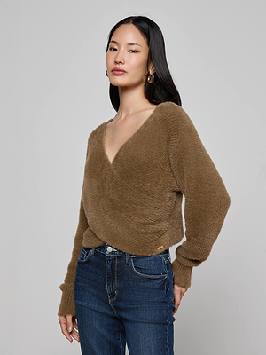 A woman with long dark hair and gold hoop earrings wears the Antonella Crossover Sweater—a cozy, chic brown fuzzy V-neck oversized pullover—with blue jeans, standing against a plain light gray background.