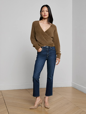 A woman stands indoors wearing the Antonella Crossover Sweater, styled with blue jeans and beige high heels. She poses with one hand in her pocket against a light gray wall and wooden floor.