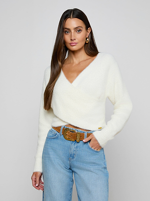 A woman with long brown hair wears the Antonella Crossover Sweater, an oversized ivory knit with a deep V-neckline, paired with light blue jeans and a brown belt. She stands facing forward against a plain light background, expression neutral.