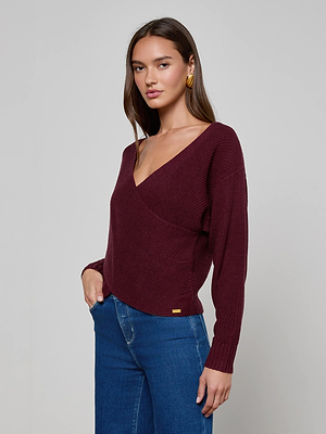 A woman with long brown hair wears the L'AGENCE Antonella Crossover Sweater—a maroon knit with a deep V-neckline—paired with blue jeans. She stands against a plain light gray background, one hand in her pocket, looking at the camera.