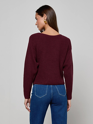A woman with long brown hair models the Antonella Crossover Sweater by L'AGENCE in maroon, featuring an oversized fit, gold hoop earrings, and stands facing away against a plain light grey background.