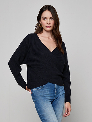 A woman with long brown hair, gold hoop earrings, and blue jeans stands against a light gray background. She wears the Antonella Crossover Sweater—an oversized knit with a deep V-neck—resting one hand on her hip and showing a neutral expression.