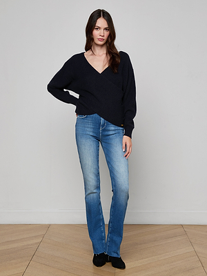 A woman with long dark hair stands against a light wall, wearing the Antonella Crossover Sweater, blue jeans, and black shoes. She has one hand in her pocket and a neutral expression.