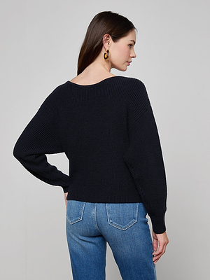 A woman with long brown hair and gold hoop earrings wears the Antonella Crossover Sweater in midnight blue with blue jeans, standing facing away from the camera against a plain light gray background.